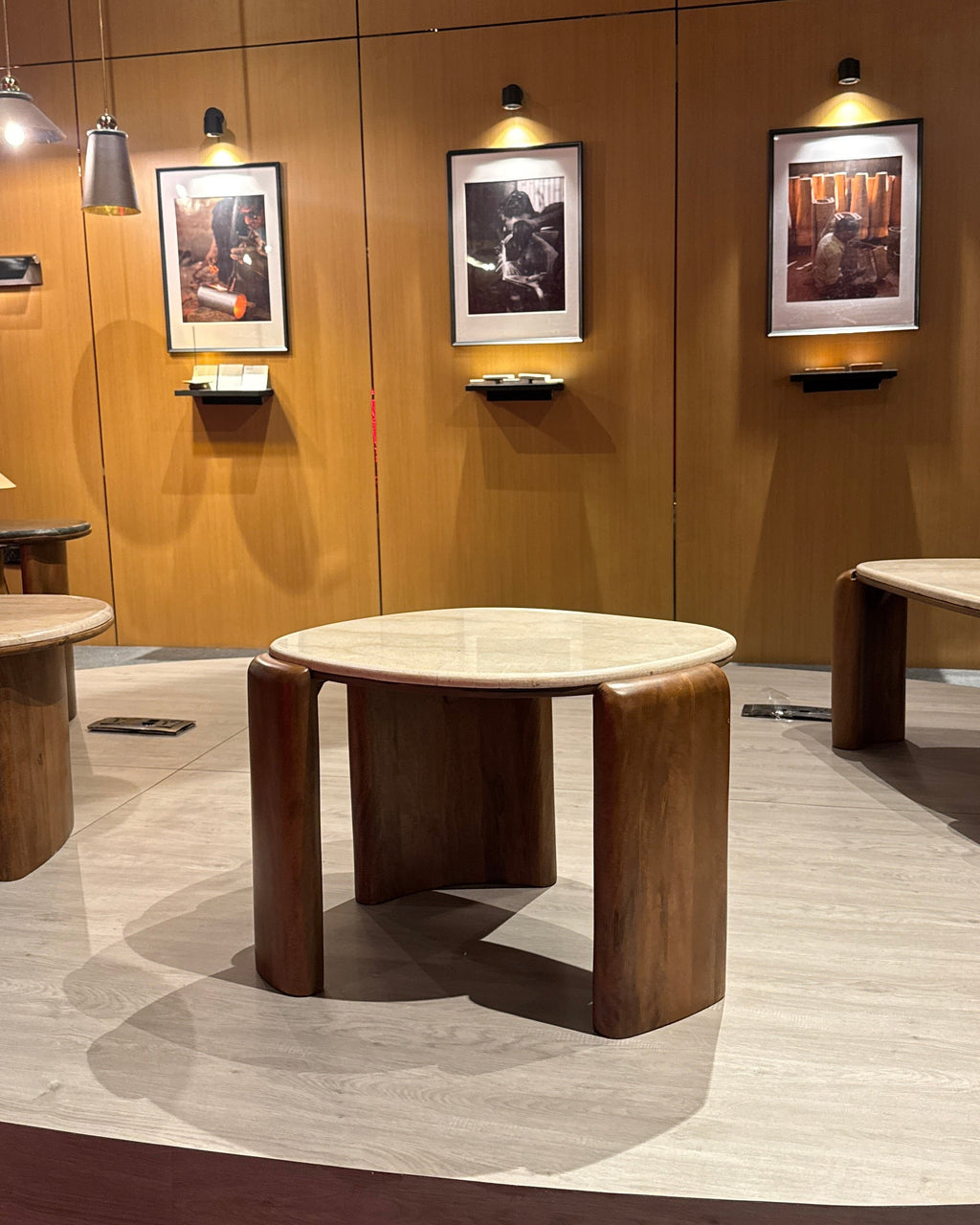 A modern round wooden table with a light stone top and thick dark wood legs, displayed in a showroom with wood-paneled walls and framed art.