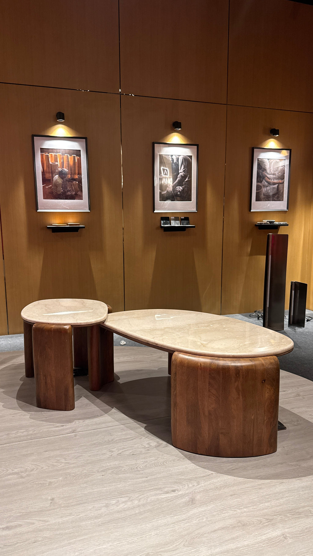 A modern coffee table set featuring a large organic-shaped oval table with a light beige marble top and dark brown wooden legs, accompanied by two matching cylindrical side tables, set in a showroom with wood-paneled walls and framed art.