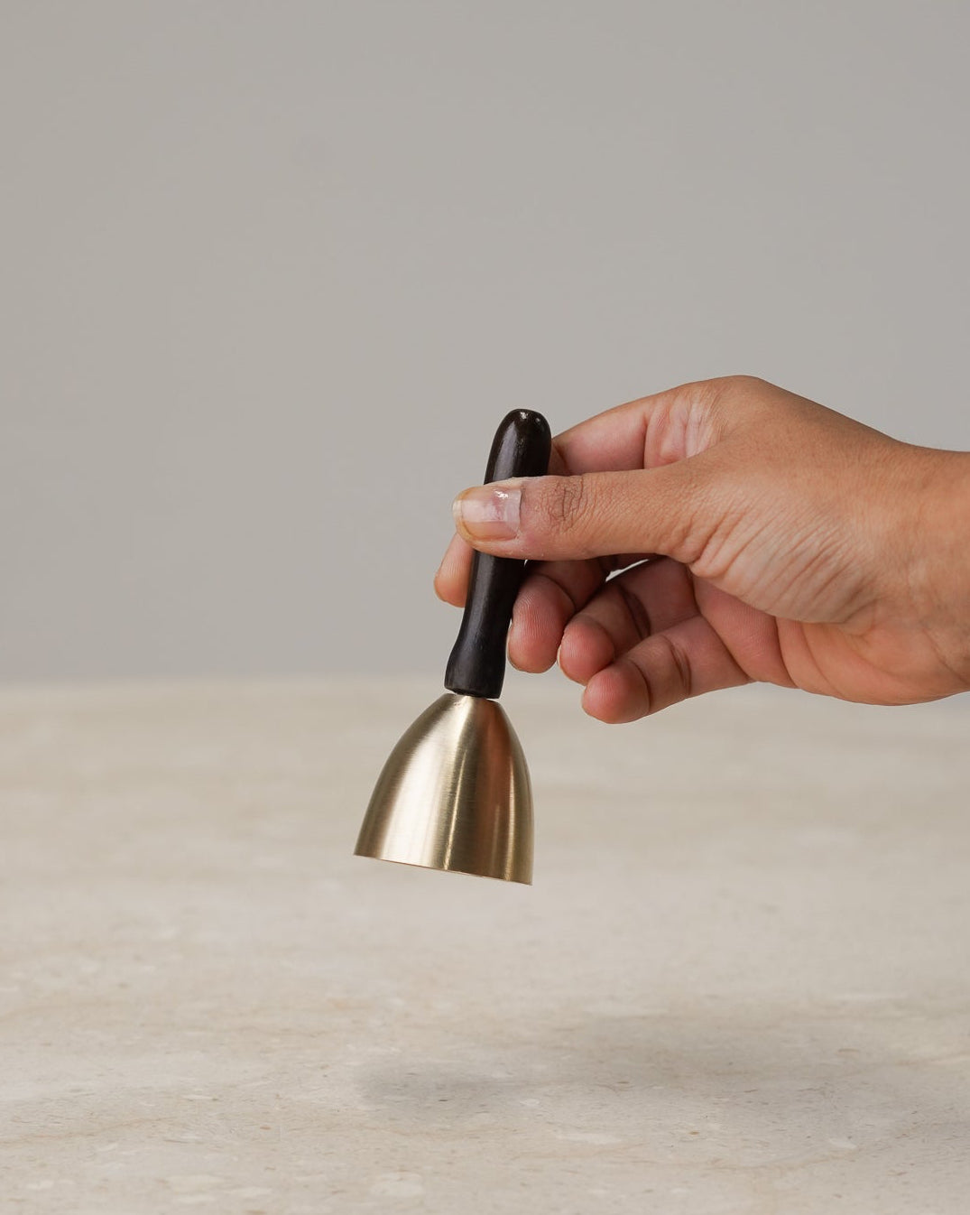 A hand holding a small brass bell-shaped tool with a black handle over a light beige marble surface against a neutral gray background.