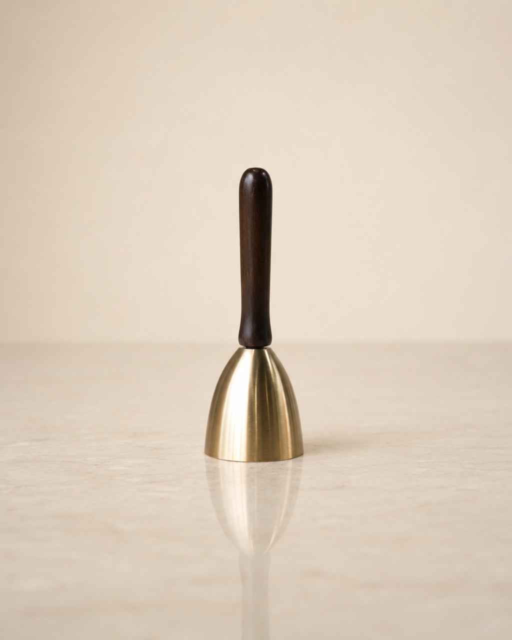 A small bell-shaped product with a polished gold-toned metal body and a dark brown wooden handle, standing upright on a light beige reflective surface against a matching neutral background.