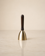 A small bell-shaped product with a polished gold-toned metal body and a dark brown wooden handle, standing upright on a light beige reflective surface against a matching neutral background.