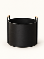 A matte black cylindrical planter or basket with two vertical side handles tipped in light natural wood, shown on a clean off-white background.