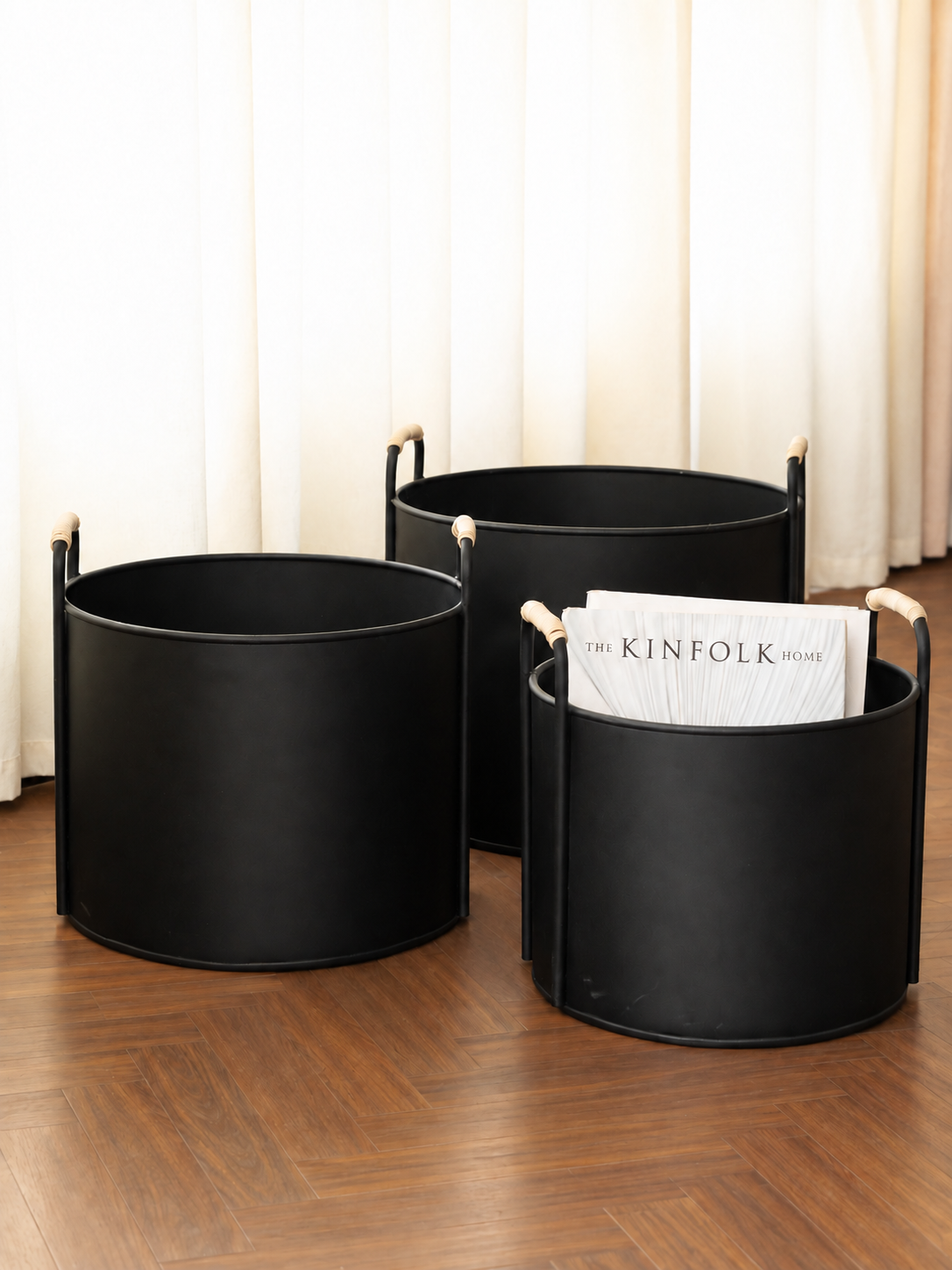 Three black cylindrical storage bins  or baskets with light wood handles arranged on a wooden floor in front of cream curtains. One bin contains a magazine.