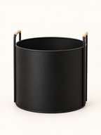 A matte black cylindrical planter or Basket with two vertical metal side supports and light natural wood handles, shown on a clean off-white background.