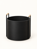 A matte black cylindrical metal planter or basket with two vertical side handles and light natural wood grips, shown on a clean white background.