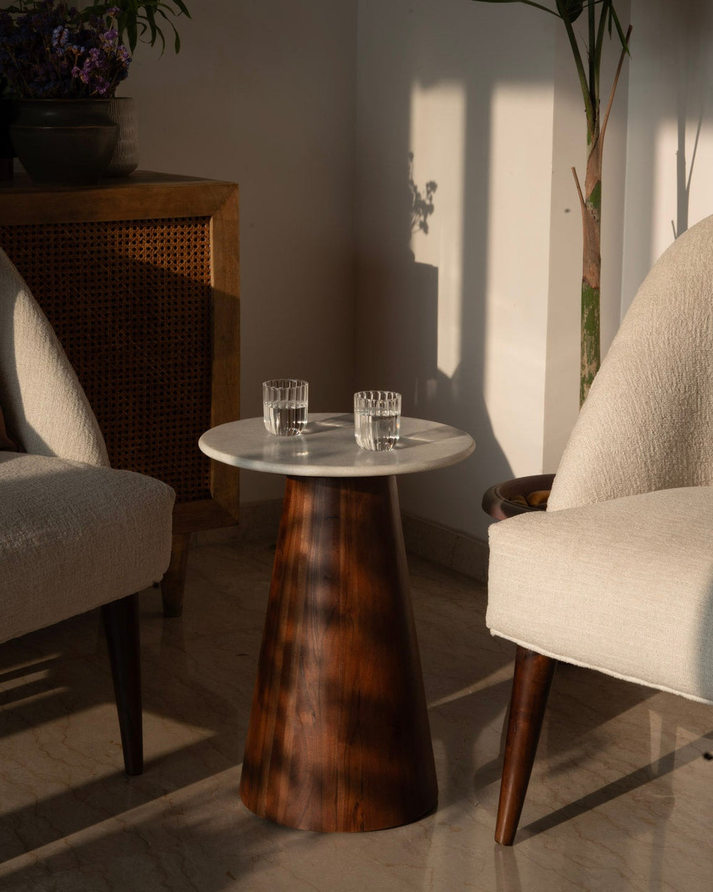 A mid-century modern side table with a round white stone top and a tapered dark brown wooden base, styled between two cream-colored armchairs in a sunlit room.