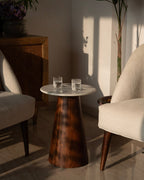 A mid-century modern side table with a round white stone top and a tapered dark brown wooden base, styled between two cream-colored armchairs in a sunlit room.