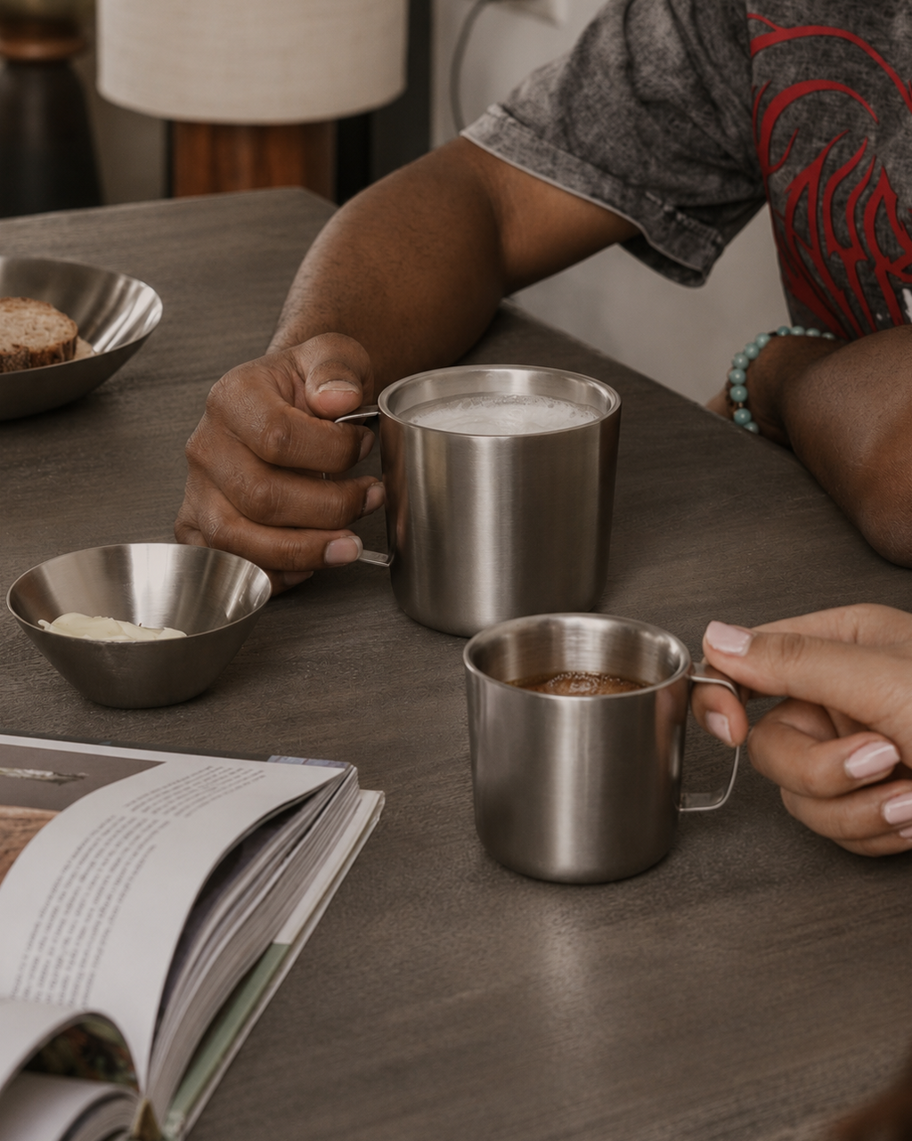 Two people holding metal cups on a wooden table with a book and bread in the background.
