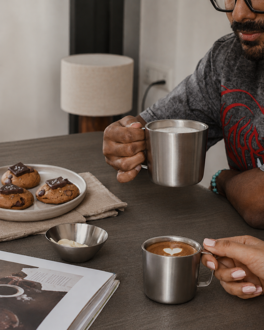 Person holding a metal mug with cookies and a magazine on a table