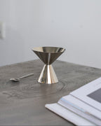 A polished Steel metal dessert coupe and a small spoon resting on a gray wooden tabletop, with a stack of white papers in the blurred foreground and a plain light gray wall behind.