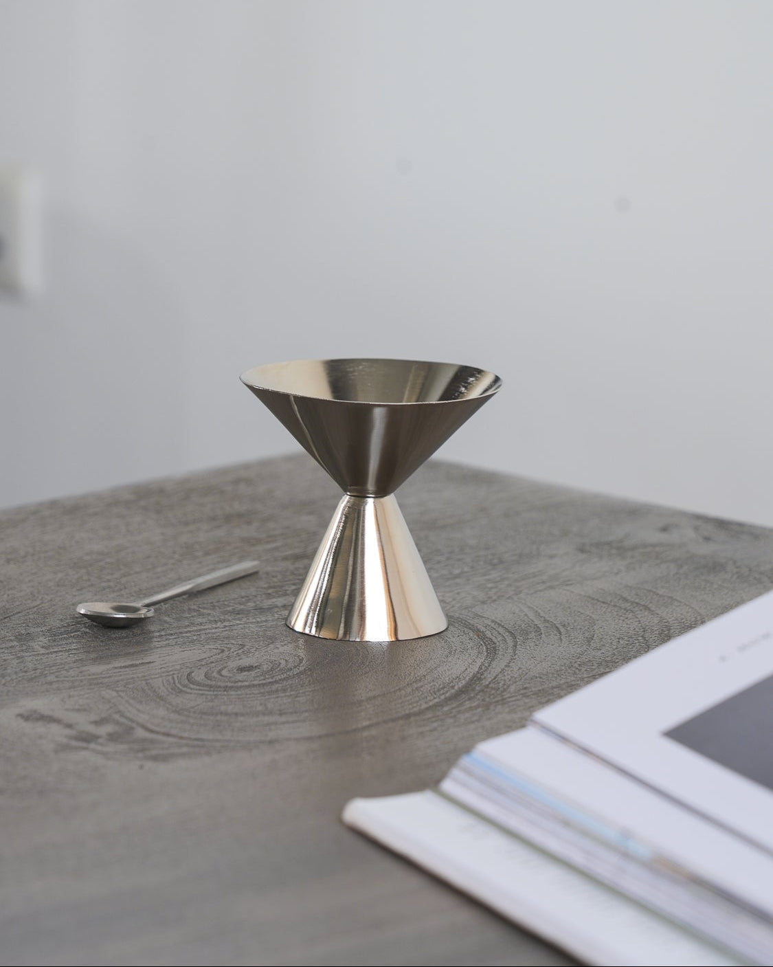 A polished Steel metal dessert coupe and a small spoon resting on a gray wooden tabletop, with a stack of white papers in the blurred foreground and a plain light gray wall behind.