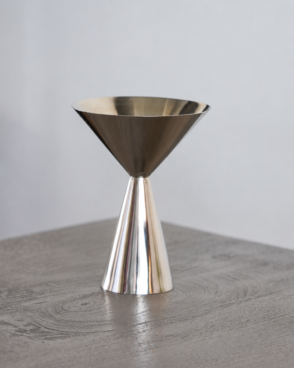 A polished metallic double jigger measuring tool with a conical base and wide funnel top, standing on a gray wooden surface against a light gray background.