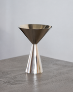 A polished metallic double jigger measuring tool with a conical base and wide funnel top, standing on a gray wooden surface against a light gray background.
