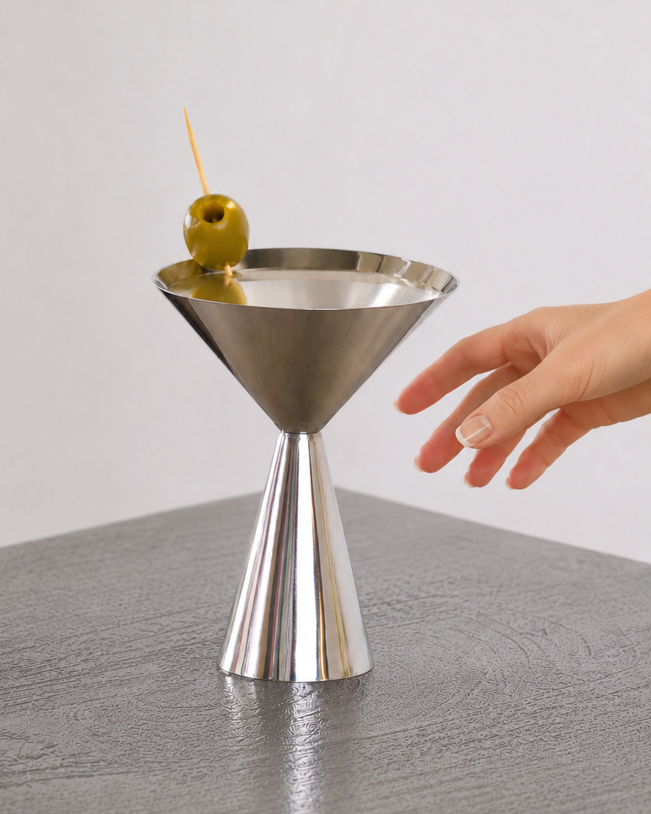 A stainless steel martini glass with a tapered base and wide conical bowl, shown on a gray tabletop with a hand reaching toward it and a green olive on a toothpick resting in the rim.