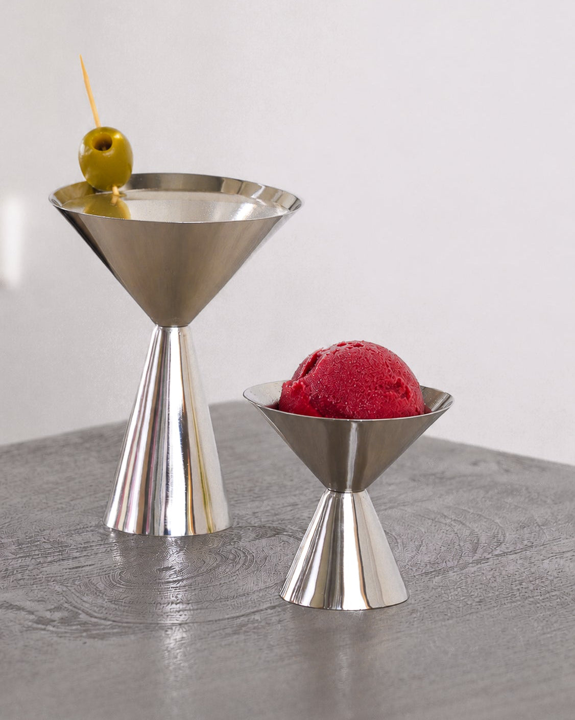 Two polished stainless steel cocktail-style serving pieces on a gray tabletop: a larger martini-shaped piece holding a green olivette, and a smaller coupe-shaped piece holding a scoop of red ice cream, against a light neutral background.