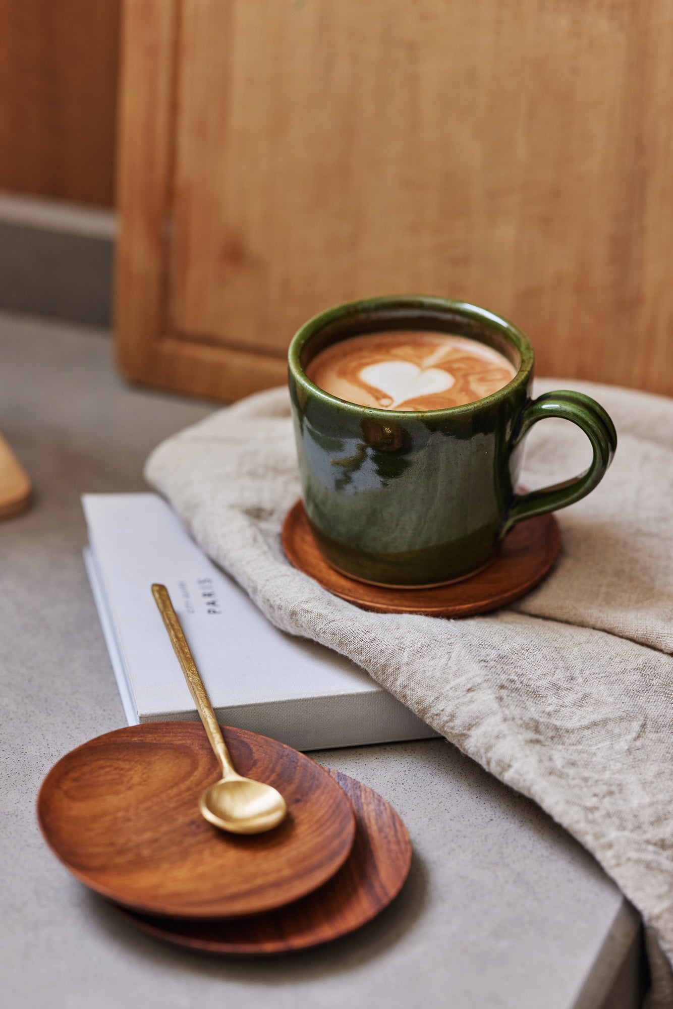 Green mug with heart-shaped latte art on a wooden coaster, placed on a book with a gold spoon beside it.