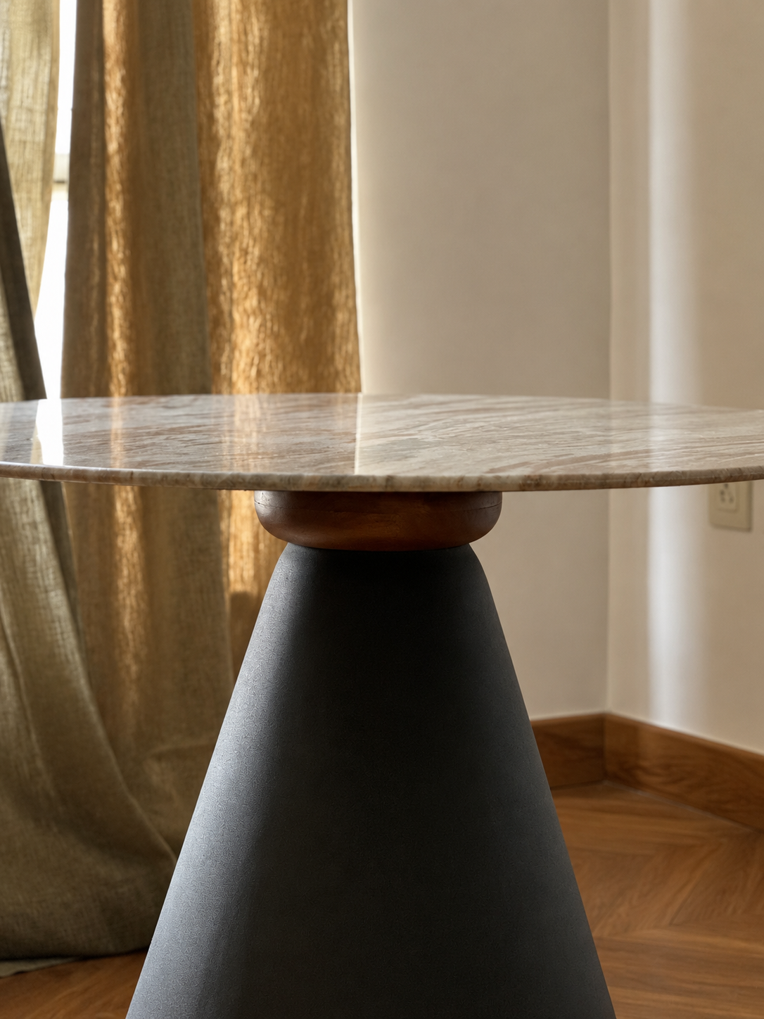 A modern round table with a light beige and white marble top, a dark charcoal gray conical base, and a small round wooden accent ring, set in a room with a gold curtain and wood floor.