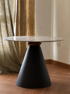 A modern round table with a light gray marble top and a matte black conical pedestal base, set in a room with a wood floor, beige walls, and textured olive-green curtains.