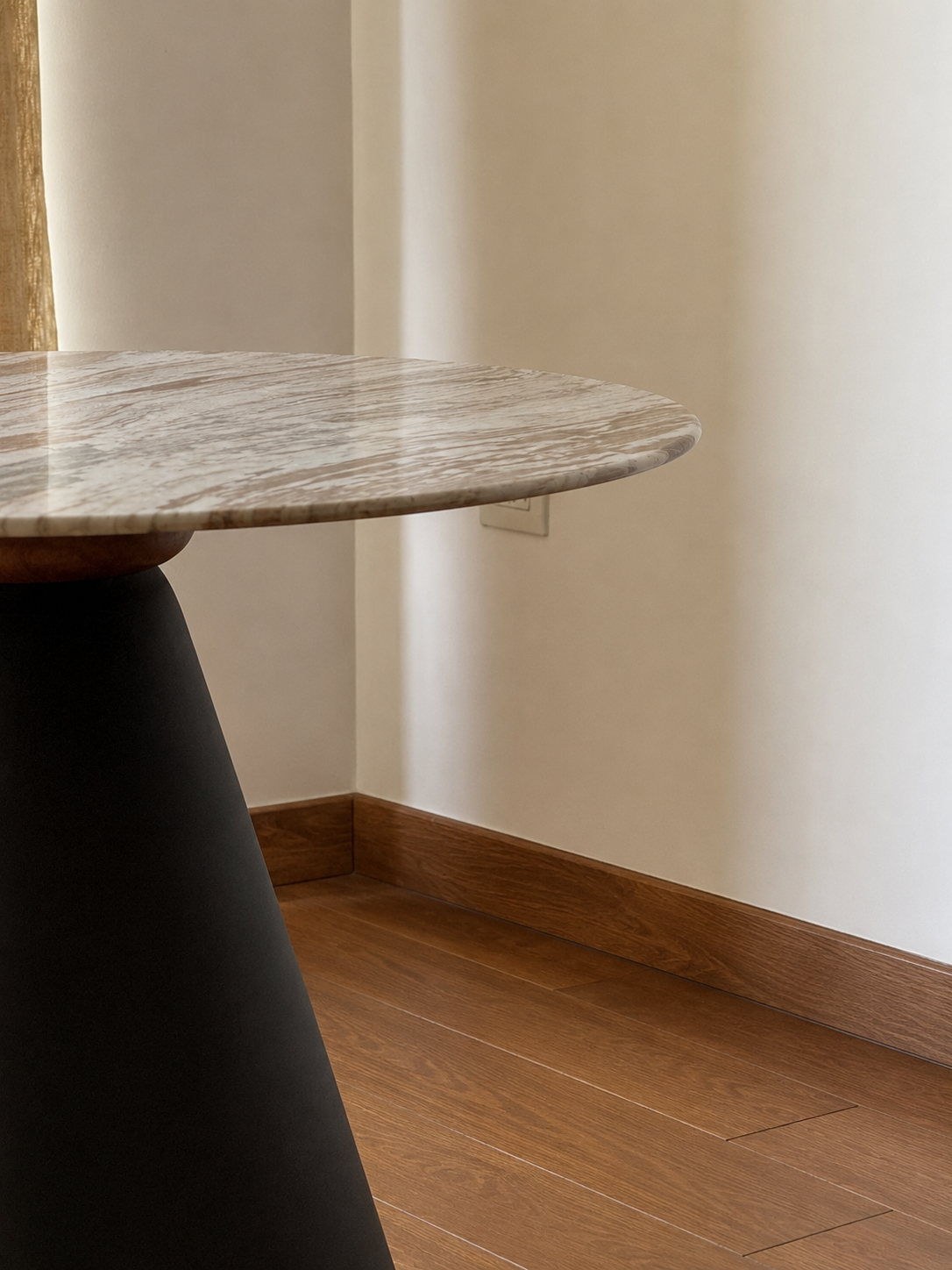 A close-up of a modern round table with a polished gray and white marble top and a dark tapered base, set in a room with beige walls and wood flooring.