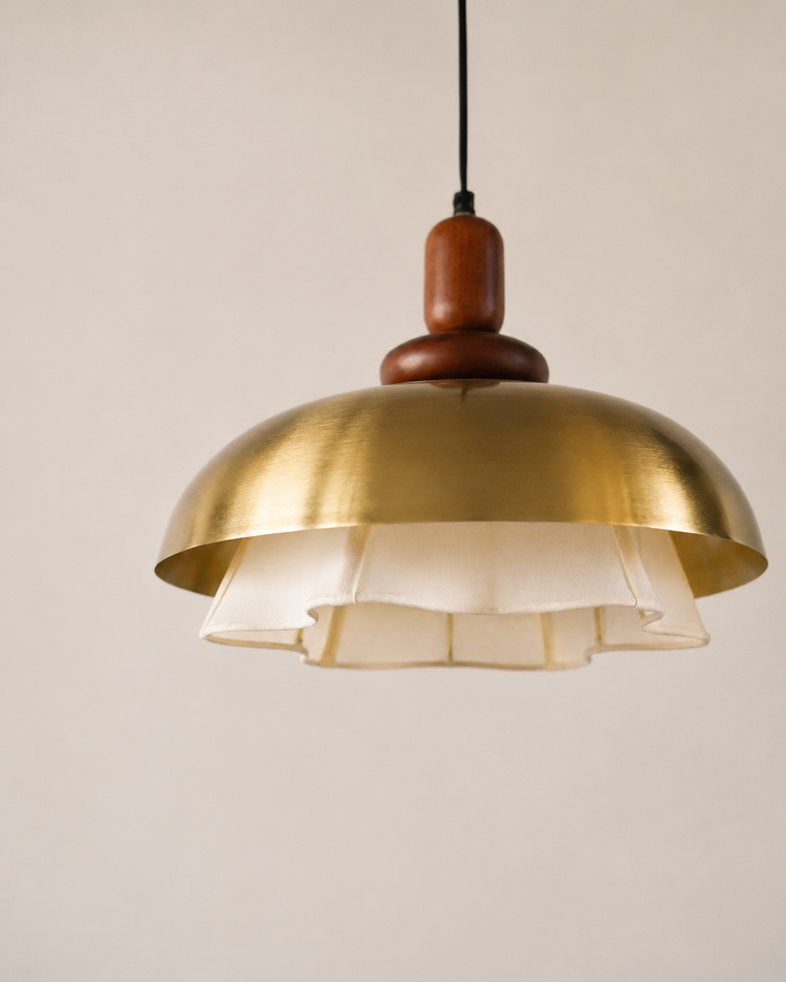 A modern pendant light with a brushed brass dome shade, a white ruffled inner shade, and a dark brown wooden neck, suspended by a black cord against a warm beige background.
