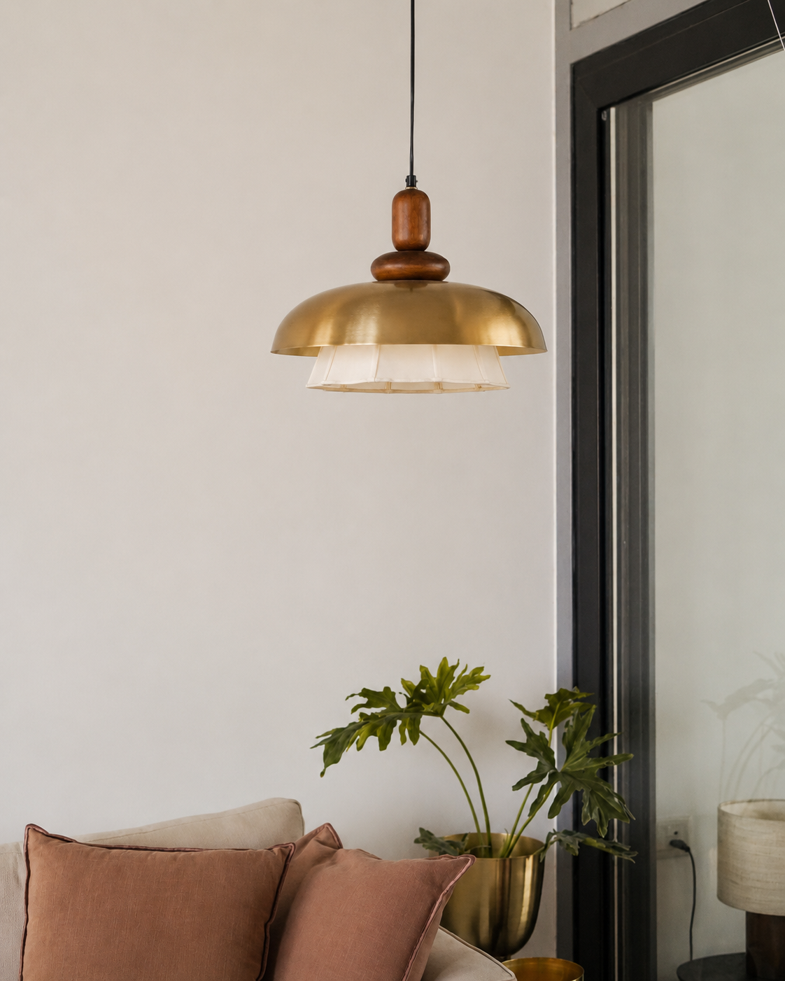 A modern pendant light with a brushed brass dome shade, a dark wood neck, and a white pleated inner shade, hanging in a minimalist room next to a black-framed glass door.