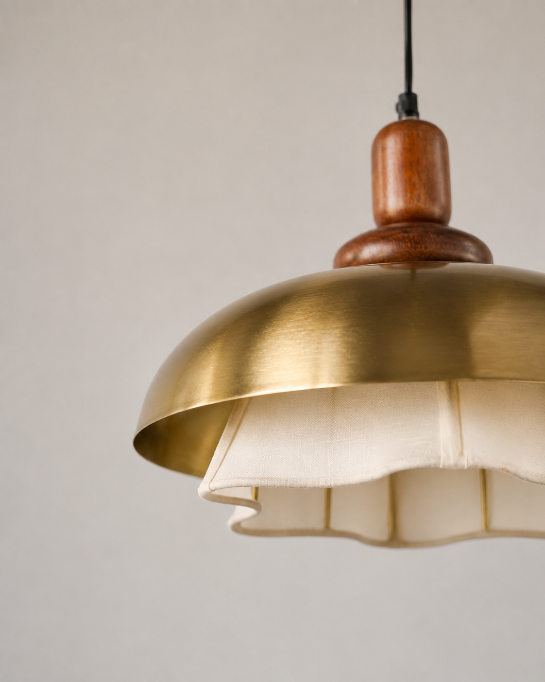 A close-up of a hanging pendant light with a brushed brass dome shade, a cream fabric inner shade, and a polished wooden finial on a black cord, set against a plain light gray background.