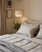 A styled bedroom scene showing a bed made with light beige quilted bedding, including a duvet cover and matching pillow shams, with a textured gray throw blanket folded at the foot. A warm pendant lamp hangs above the bed, and framed wall art and curtains are visible in the softly lit room.