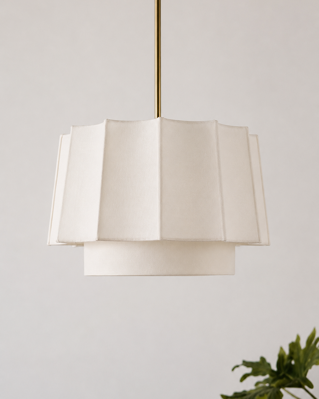 A modern tiered pendant light with a cream-colored pleated fabric shade and a slim brass suspension rod, shown against a neutral light gray wall with a green plant leaf in the lower right corner.