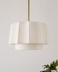 A modern tiered pendant light with a cream-colored pleated fabric shade and a slim brass suspension rod, shown against a neutral light gray wall with a green plant leaf in the lower right corner.