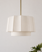 A modern tiered pendant light with a cream-colored pleated fabric shade and a slim brass suspension rod, shown against a neutral light gray wall with a green plant leaf in the lower right corner.