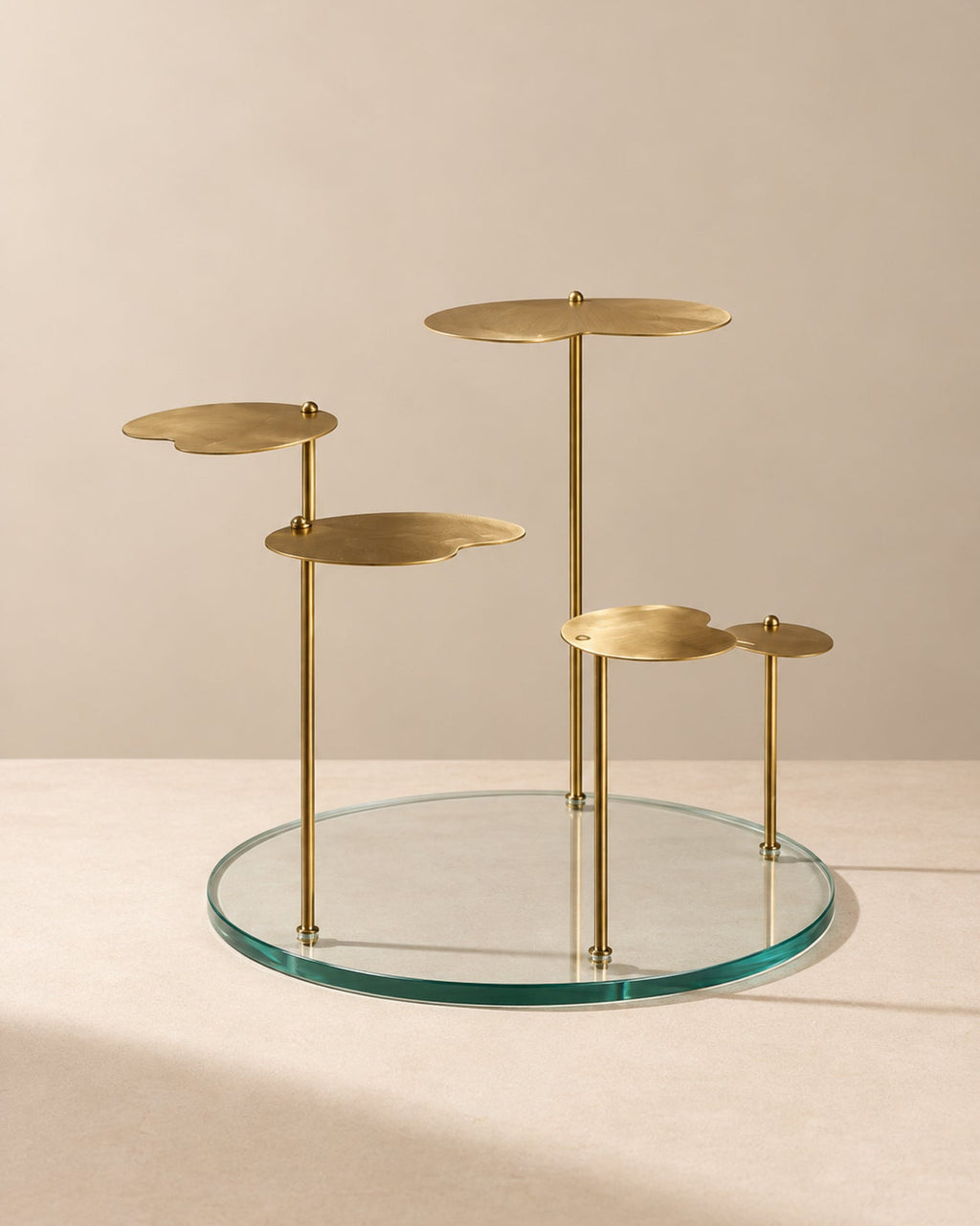 A modern brass and glass side table featuring a circular clear glass base, four slim brass vertical posts, and four organic-shaped brass platforms at varying heights, set against a warm beige background.