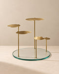 A modern brass and glass side table featuring a circular clear glass base, four slim brass vertical posts, and four organic-shaped brass platforms at varying heights, set against a warm beige background.