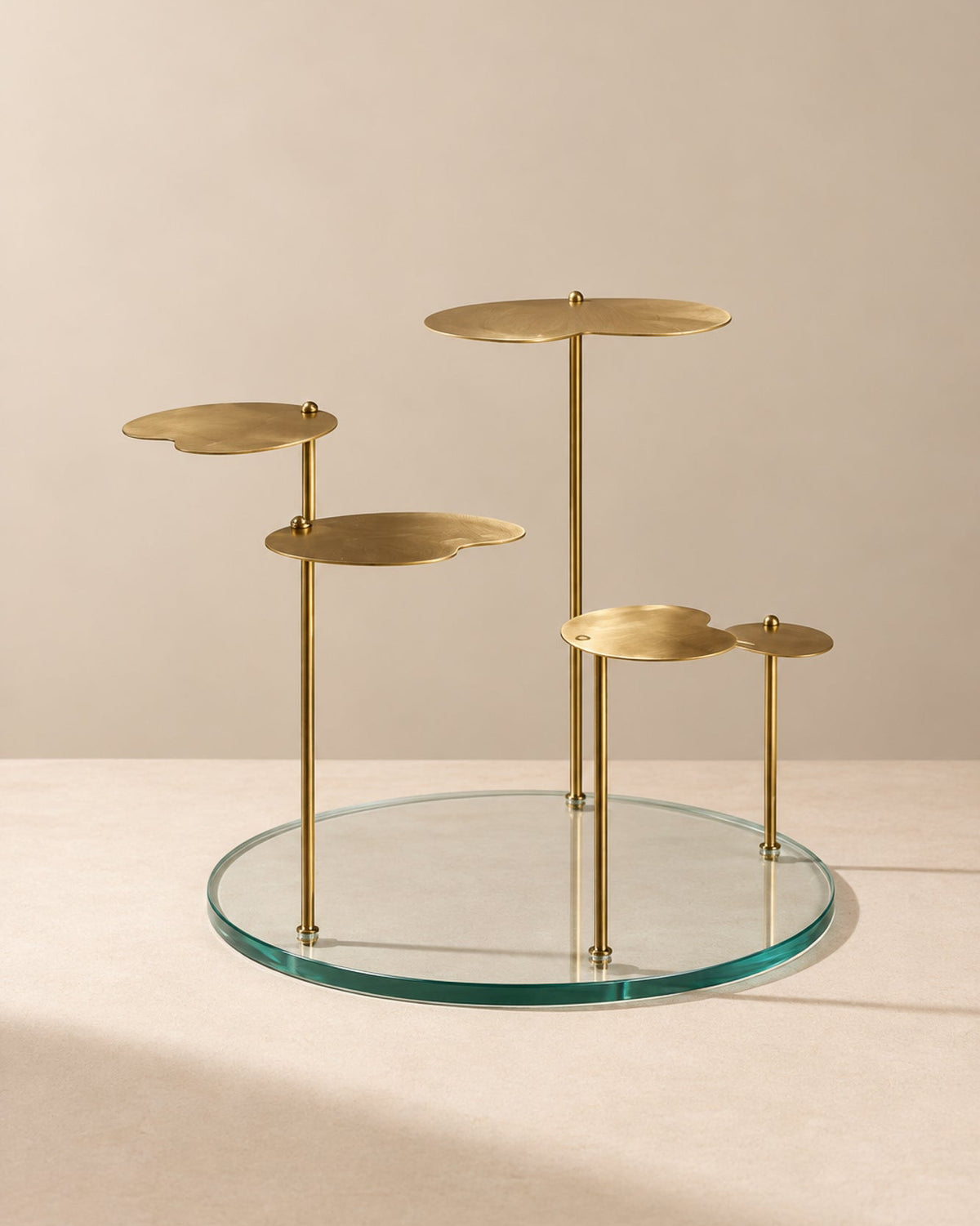 A modern brass and glass side table featuring a circular clear glass base, four slim brass vertical posts, and four organic-shaped brass platforms at varying heights, set against a warm beige background.