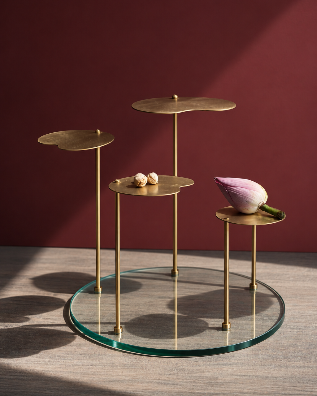 Three-tier brass side table set with a clear glass circular base, styled with a pink lotus flower and small decorative objects against a dark red background.