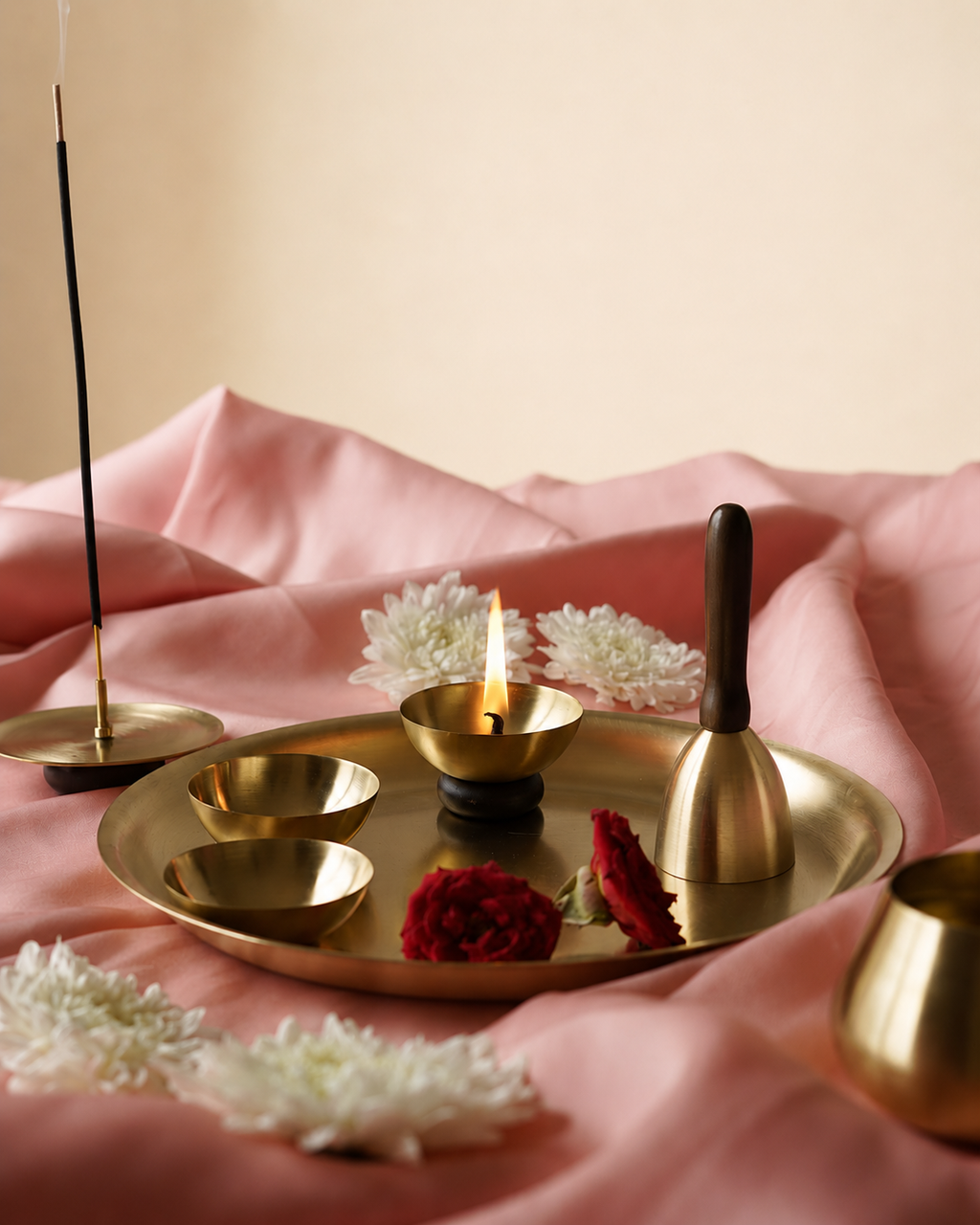 A brass ritual set arranged on draped pink fabric, including a tray, small bowls, a bell with a dark handle, a tall incense stick holder, a lit oil lamp, and white flowers.