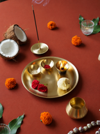 A styled flat-lay of a brass ritual altar (pradaksha) featuring a round gold tray with small bowls, a burning incense holder, and a lit oil lamp, surrounded by decorative elements like marigolds, coconut, and leaves on a terracotta background.