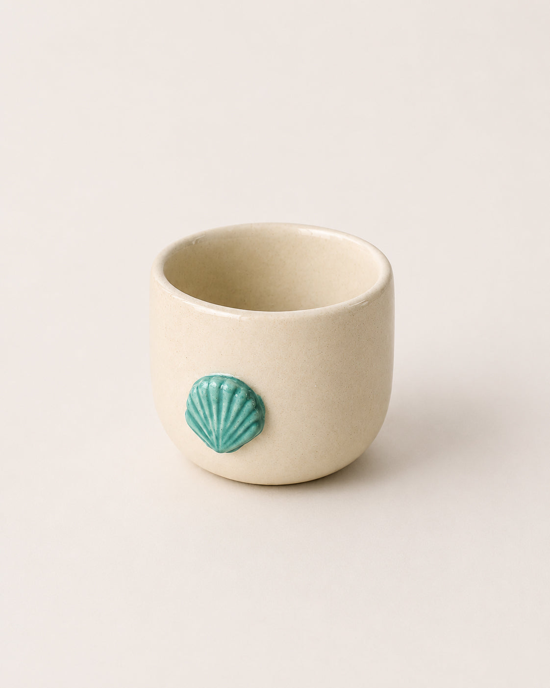 A small beige ceramic cup with a glossy turquoise shell motif on the front, set against a solid off-white background.