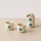 Four small ceramic cups in a warm cream color, each decorated with a raised teal shell motif, arranged on a light wood surface against a soft off-white background.