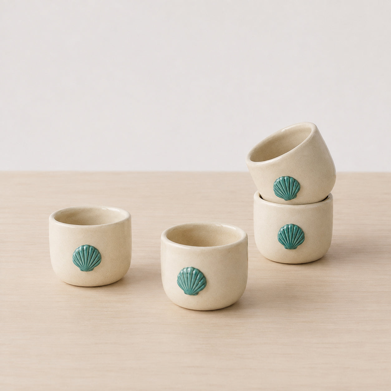 Four small ceramic cups in a warm cream color, each decorated with a raised teal shell motif, arranged on a light wood surface against a soft off-white background.