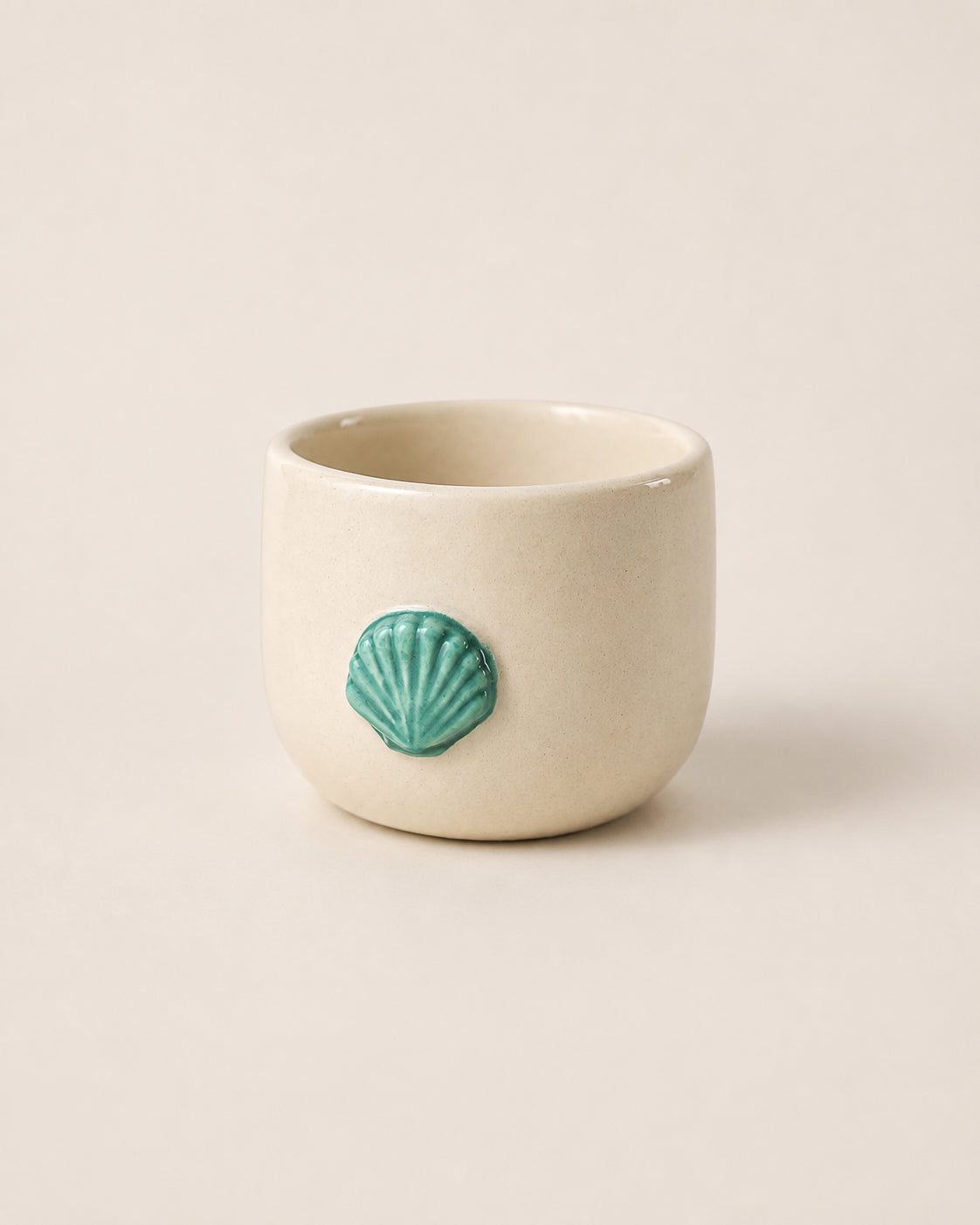 A small cream-colored ceramic cup with a textured finish and a raised turquoise-green seashell accent on the front, set against a solid light beige background.