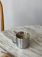 Stainless steel mug with coffee on a marble surface next to pastries