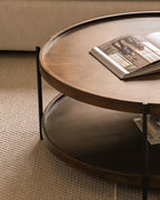 A close-up view of a modern round wooden coffee table with a lower shelf, set on a textured beige carpet.