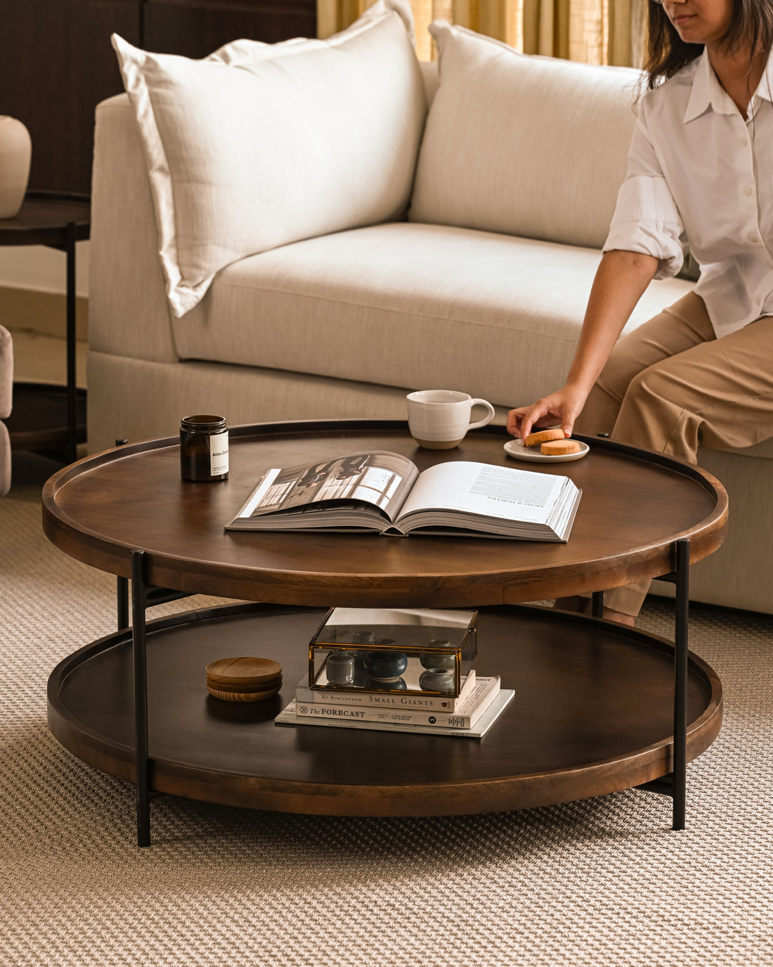 Round wood coffee table with metal legs, open shelf, and styled with books, mug, and decor in a cozy living room.