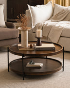 Round small wooden coffee table with metal frame and lower shelf in living room with a beige sofa in the background.