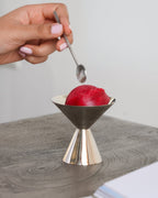 A hand with pale pink manicured nails holds a small metal spoon above a metallic cone-shaped dessert holder containing a red frozen dessert, set on a gray tabletop against a light neutral background.