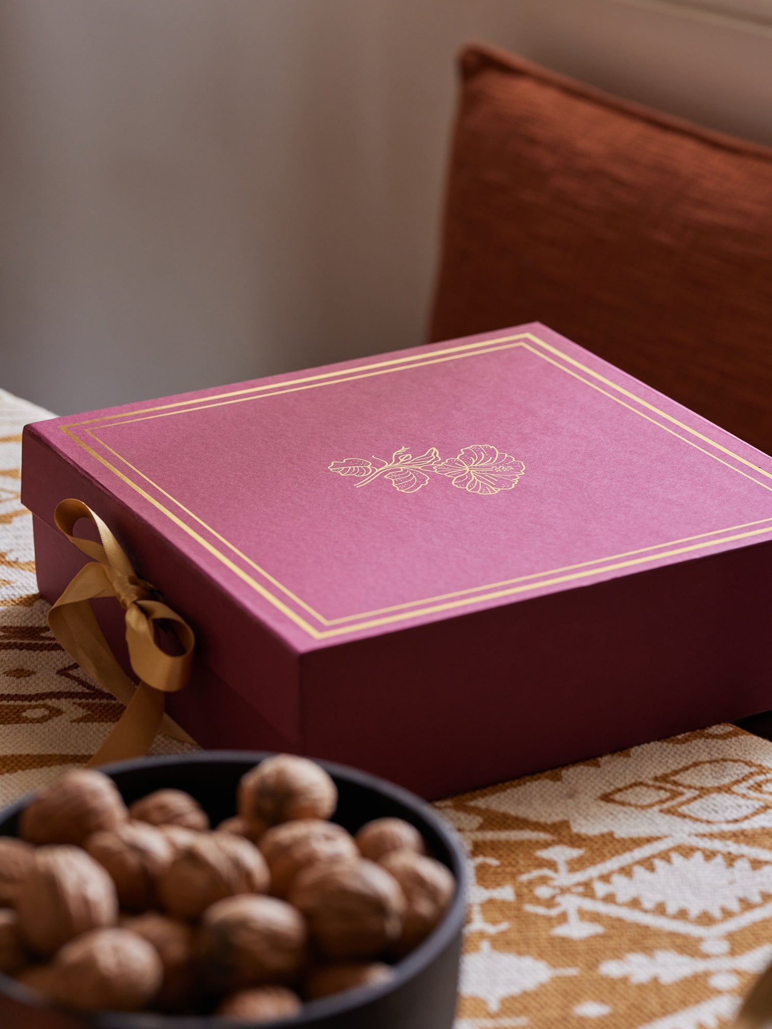A rectangular magenta gift box with gold foil borders and a gold ribbon bow, placed on a patterned tablecloth next to a bowl of walnuts, with a brown cushion in the background.
