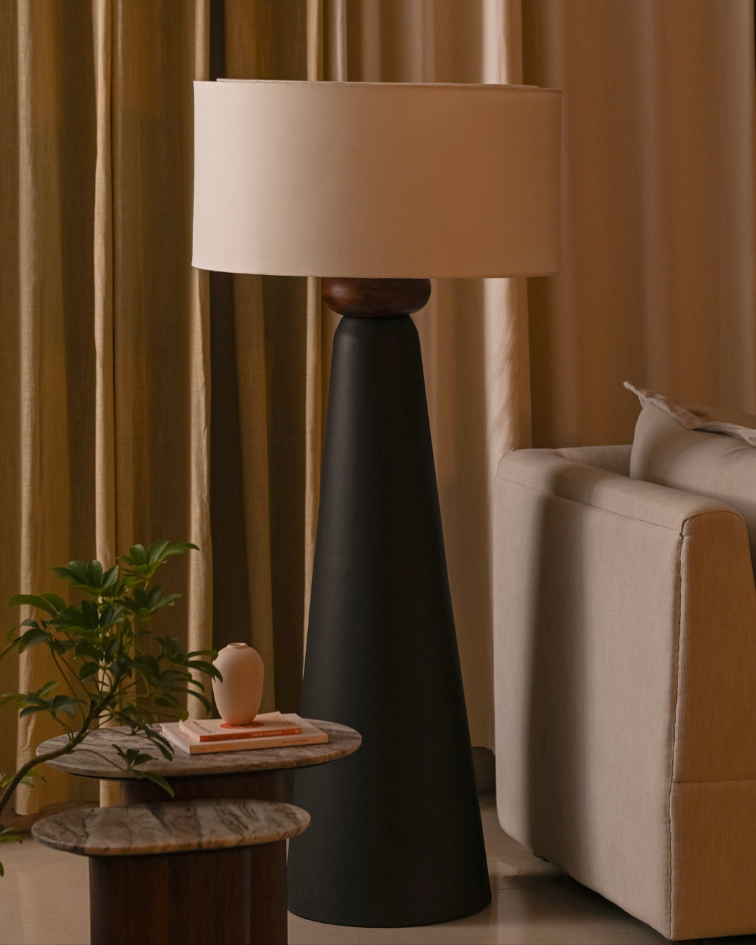 Floor lamp with a beige shade in a living room setting with curtains and a sofa.